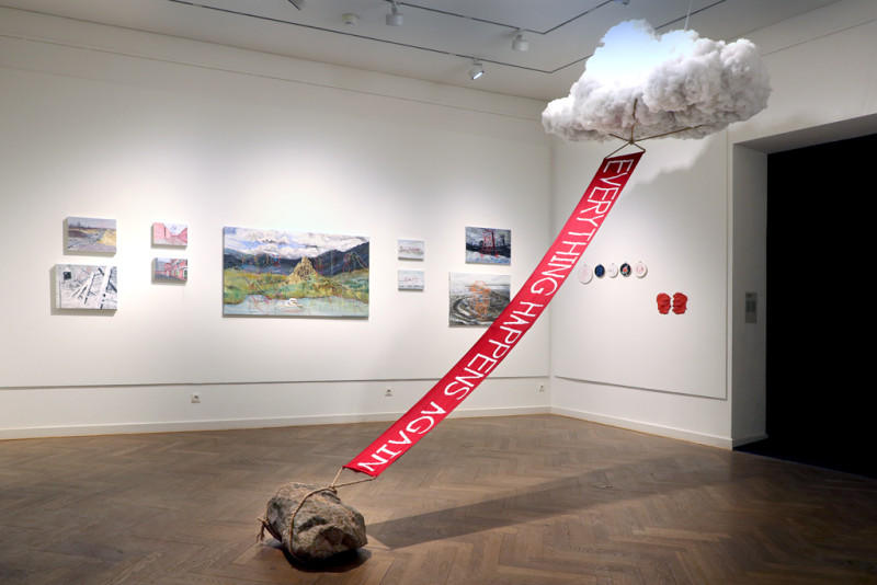 Rauminstallation. Von der Decke hängt eine Wolke, die durch eine rote Fahne mit einem Stein, am Boden verbunden ist.