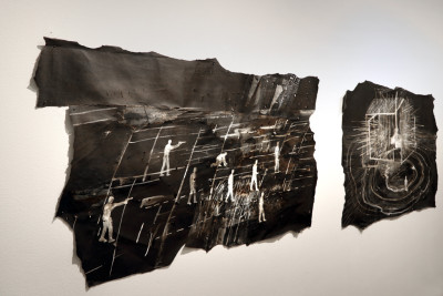 Two bitumen on the wall. On one are depicted armed people, which is located in a field. On the other bitumen a house with barbed wire. The images are abstract and painted with white oil pastels and acrylic paints.