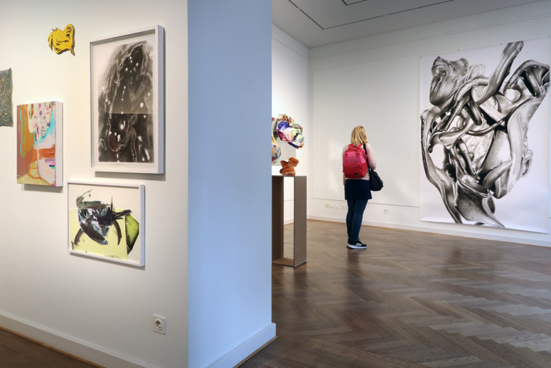 Links sind mehrere kleine Bilder an der Wand. Rechts: Blick in den zweiten Raum. Dort steht eine Besucherin vor der großformatigen Zeichnung von Peter Hock.