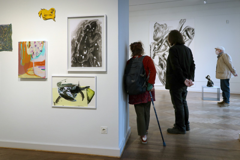 Ausstellungsansicht: fünf kleinformatige Bilder hängen links an der Wand, rechts Blick in den zweiten Ausstellungsraum mit Besuchenden der Ausstellung.