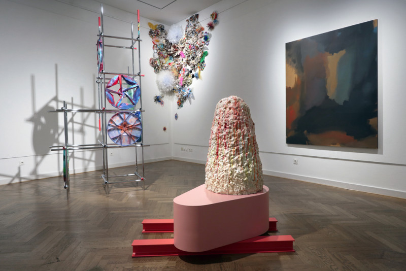 Es ist eine Raumansicht zu sehen mit einem rosa Objekt im Vordergrund, rechts dahinter eine Installation aus Metallstangen und Autofelgen sowie an der Decke Plastikblumen. An der rechten Wand hängt ein abstraktes Bild mit dunklen Farben.