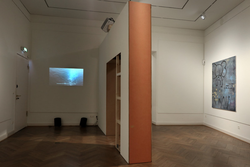 Zu sehen ist ein durch ein Raumelement zweigeteilert Raum. Links gibt es eine Videoprojektion an der Wand, rechts hängt ein graues Gemälde an der Wand.