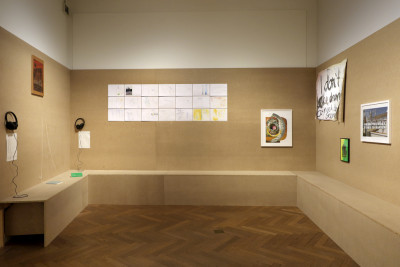 Eine Raumansicht, wo den Wänden eine weitere braune Wand vorgelagert ist mit einer Sitzbank. An der brauen Wandfläche sind verschiedene Kunstwerke angebracht sowie Kopfhörer.