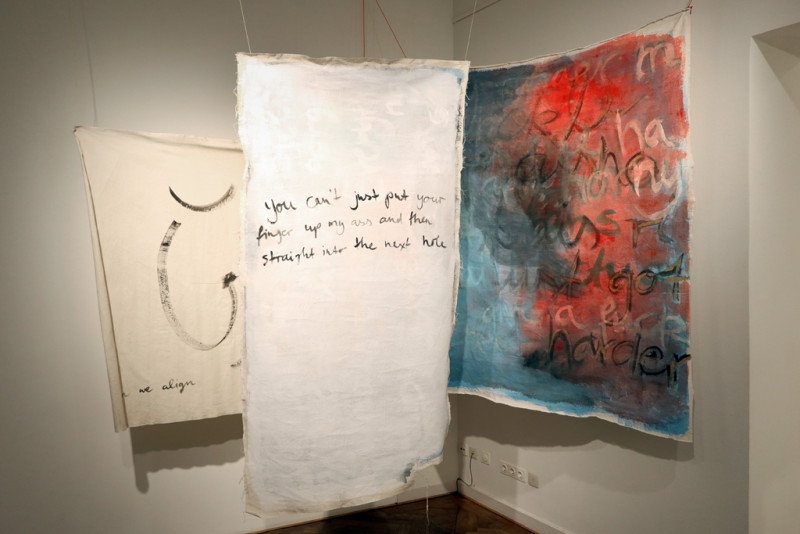 Es sind drei von der Decke hängende Leinwände zu sehen, mit bunten Farben und Text beschrieben.