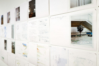 Auf einer Wand sind Reihen von A4 Blättern mit Nägeln angebracht, die handschriftliche Texte zeigen sowie Fotos.