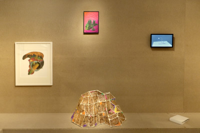 Es ist eine braune Wand mit Sitzbank zu sehen. Darauf sind gerahmte Collagen sowie ein schwarzer kleiner Monitor angebracht. Auf der Sitzbank steht ein braunes Objekt sowie ein Buch.