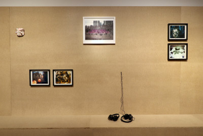 Es ist eine braune Wand mit Sitzbank zu sehen. Darauf sind gerahmte Collagen angebracht sowie ein Keramikobjekt und Kopfhörer.