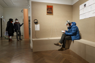 Im Vordergrund ist eine braune Wand mit Sitzbank zu sehen. Dort sitzt ein Besucher mit Kopfhörern. Im hintergrund sind weitere Besucher:innen im Ausstellungsraum zu sehen.