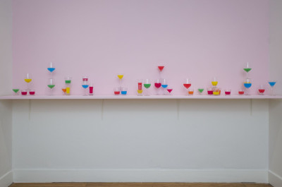 Auf einer Regalleiste an der Wand stehen Gläser in verschiedenen Formen, gefüllt mit bunten Flüssigkeiten