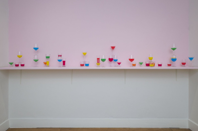 Auf einer Regalleiste an der Wand stehen Gläser in verschiedenen Formen, gefüllt mit bunten Flüssigkeiten
