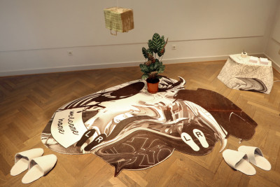 Room instrallation consisting of a brown and white carpet, around it are slippers and a small table. A golden bag hangs from the ceiling.