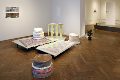 View of the room. Thre is a meditarranean room installation with carpet and beanbags (made of various materials) on the floor. Cardboard columns and plants stand alongside. In the back room you can see a door on the pedestal.