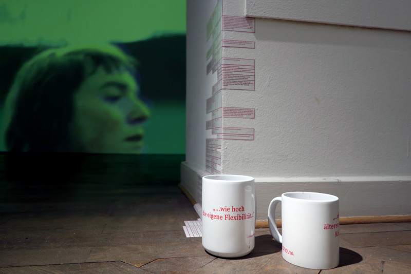 View of the room from below. In the foreground on the floor are cups with red writing. On the corner of the wall there are strips of paper with red writing. In the room behind, a green picture (Film clip) is projected on the wall with the beamer.