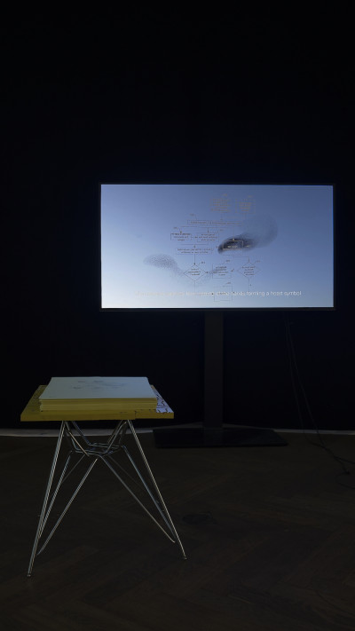 Dark view of the room. There is a metal table with a yellow tabletop and a stack of of papers on it. A blue glowing screen shows an explanatory graphic.