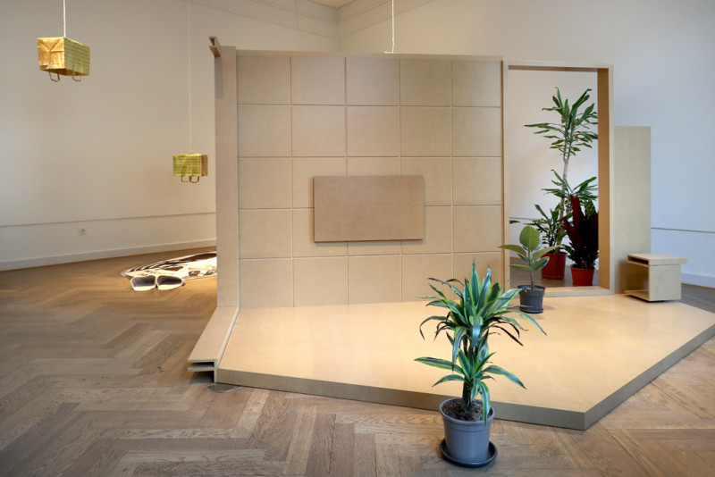 Raumansicht. Die Installtion zeigt einen mediterranen Raum aus Holz, Pflanzen stehen herum, goldene Tüten hängen von der Decke.