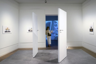 Room view. Two white doors stand on a pedestal. Pictures hang on the walls, in the background the other room is dark. There something is projected on the wall with a beamer.