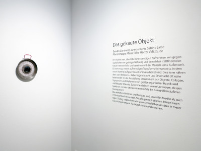 View of a corner of a room, on the left wall is an eye-shaped object made from a paella pan, protractors and sewing silk, on the right wall is the exhibition text for the exhibition 