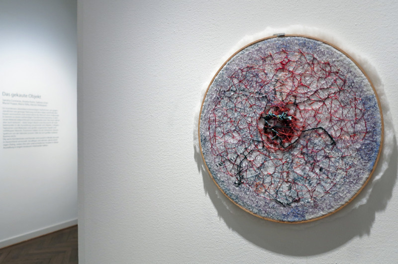 A red and blue embroidery in a round wooden embroidery frame, the shape resembles a network of nerves or concentric blood vessels.