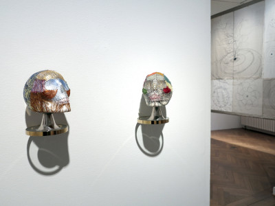 On the wall in the foreground are two skull-like sculptures wrapped in colored sewing silk, while a translucent cotton hanging with black embroidery can be seen in the background through the passageway next to it.