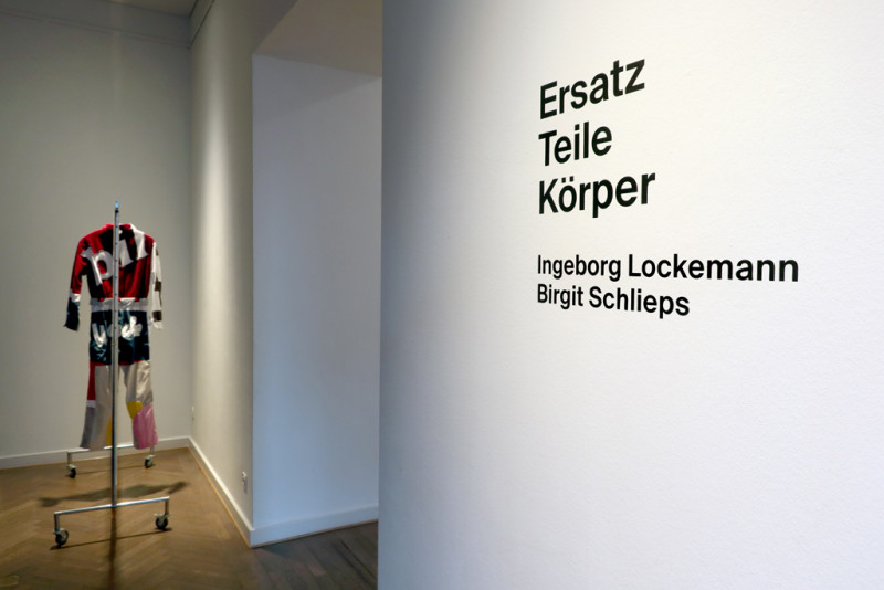 Im Vordergrund: Titel und Künsterinnen der Ausstellung, Im Hintergrund: Bunter Werkstattanzug