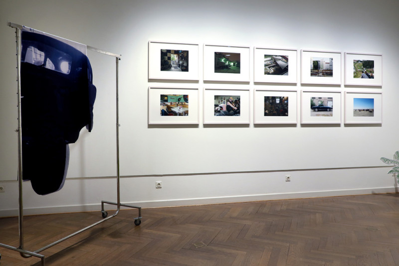 Ein Kleiderständer steht im Raum, daran befestigt ist die Installation. An der Wand hängen eingerahmte Fotos.