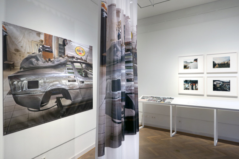 On the left on the wall: large poster with a car body and a textile with the same image in front of it, on the right: photographs of various car repair stores
