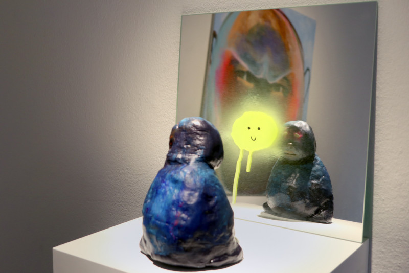 A blue little sad sculpture sits on a pedestal. The sculpture is looking into a mirror that is in front of it. On the mirror is a yellow smiling face.