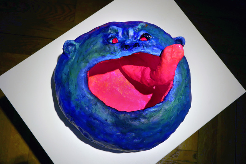 Eine Gesicht-Skulptur, in blau und pink steht auf einem Podest.