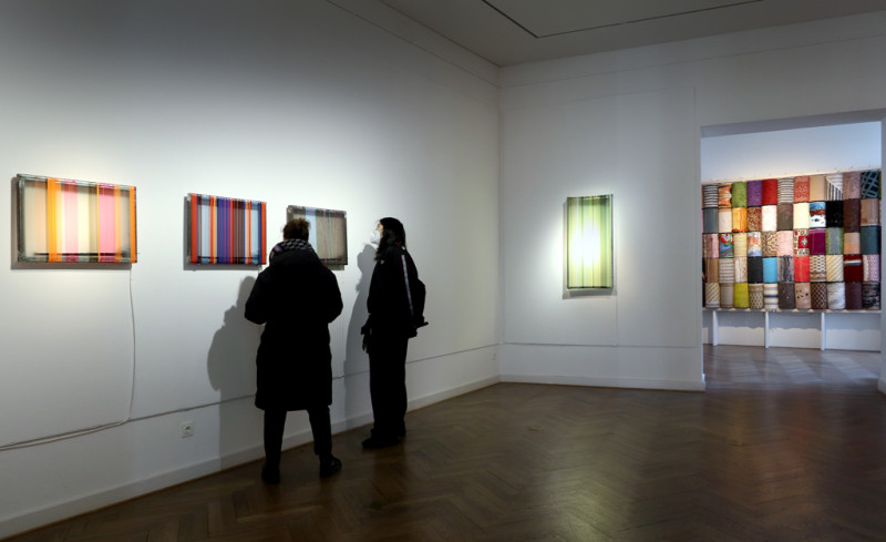 Zwei Besucherinnen stehen an der linken Seite vor drei kleinformatigen Bildern an der Wand, die aus Angelschnürgen bestehen.