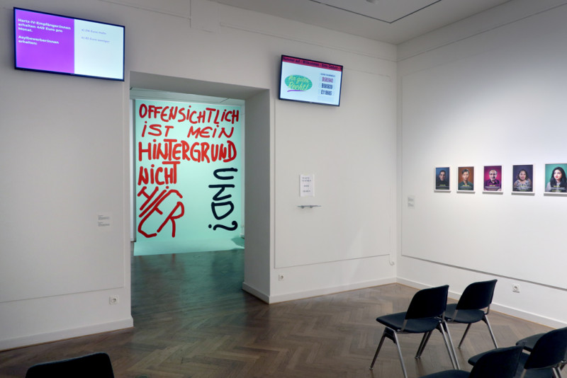View through the premises. There are highmounted luminous screens and chairs , portrait pictures and the blue poster with red and black lettering.