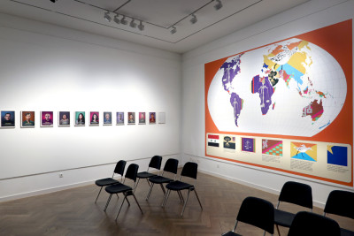 Blick in den Raum. Es stehen Stühle in Reihen aufgestellt, an den Wänden hängen 12 Portrait-Bilder und ein großes Plakat, darauf ist die Erde mit ihren Kontinenten abgebildet. die Kontinente und Länder sind neu kategorisiert und aufgeteilt.