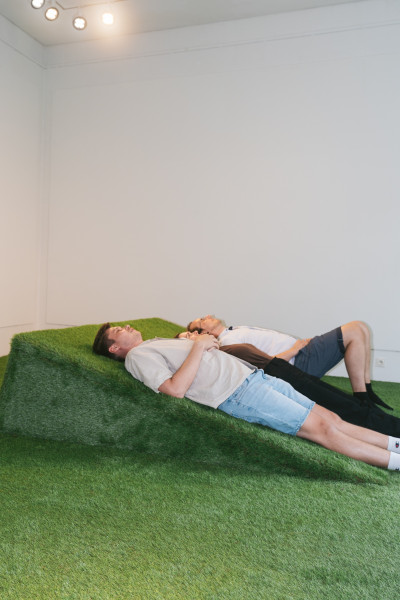 Zwei Personen liegen entspannt auf einer grünen Kunstrasen-Installation in einem weißen Galerieraum