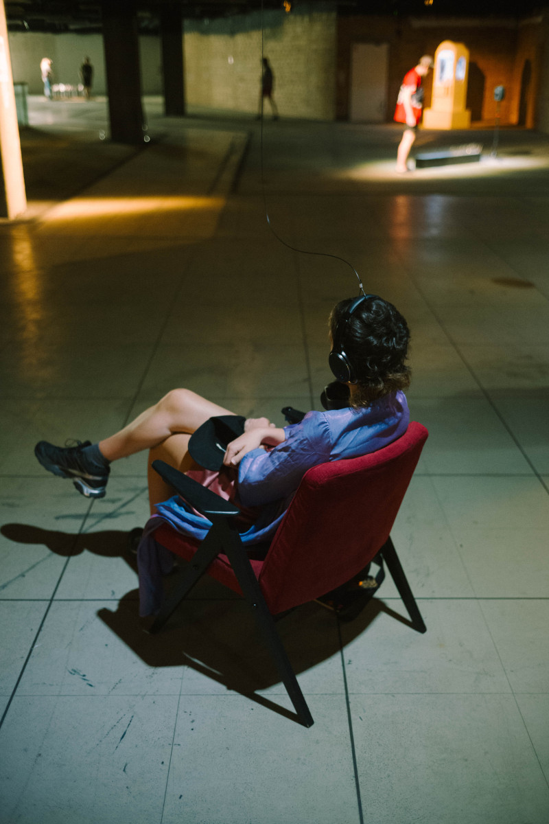 Eine Person sitzt in einem Ausstellungsraum auf einem Stuhl. Der Stuhl gehört zu dem Kunstwerk 