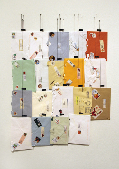 19 Briefe in verschiedenfarbigen Umschlägen hängen in einem Block an Schnüren befestigt an der Wand.