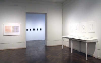 Links sind eine rote und eine blaue Zeichnungen zu sehen. In der Mitte hängen fünf kleine rechteckige schwarze 3D-Drucke an der Wand. Auf der rechten Seite hängen vier Schwarz-Weiß-Fotografien von Gebetsketten.