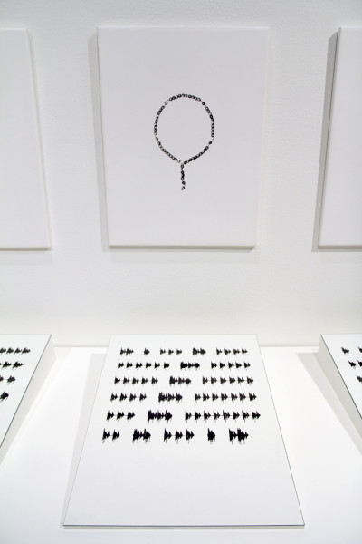 Eine Schwarz-Weiß-Fotografie von einem Gebetskranz hängt an der Wand, davor liegt eine Tuschezeichnung mit einer sogenannten Phonokalligrafie.