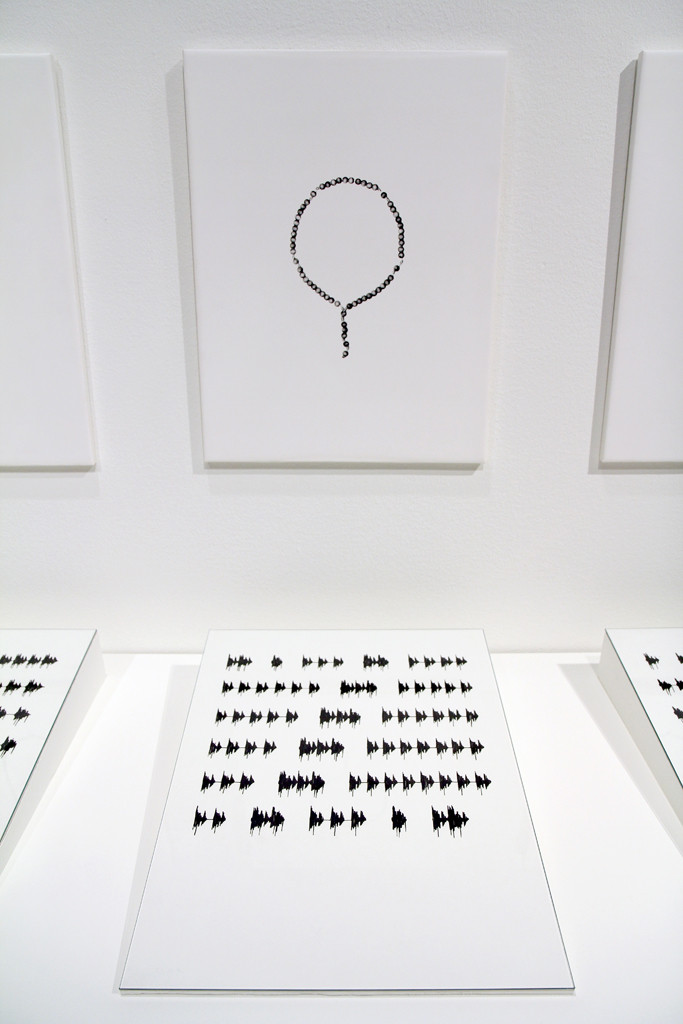 Eine Schwarz-Weiß-Fotografie von einem Gebetskranz hängt an der Wand, davor liegt eine Tuschezeichnung mit einer sogenannten Phonokalligrafie.