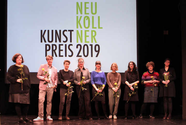 Neun Menschen stehen mit Blumen in der Hand auf einer Bühne vor einer Projektion der Schrift Neuköllner Kunstpreis 2019.