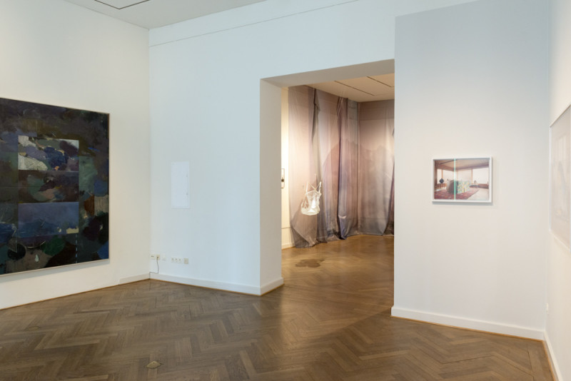 Links hängt ein in dunklen Tönen gehaltenes abstraktes Gemälde. Im mitttleren Raum sind Gurtzeug und Fallschirmseide an der Wand und im Raum angebracht. An der rechten Wand ist eine Fotografie von einem Wohnzimmer zu erkennen.