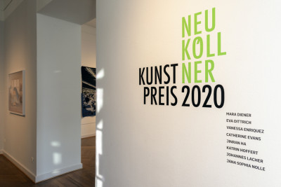 Das Logo zum Neuköllner Kunstpreis 2020 ist in grüner und schwarzer Schrift auf der Wandfläche in der Ausstellung zu sehen.