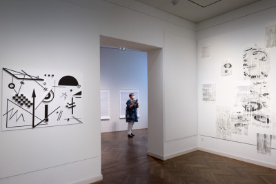 Auf der rechten Seite ist ein absraktes schwarz weißes Bild an der Wand und auf der rechten Seite ist eine schwarz weiße Papierarbeit an der Wand. In der Mitte des Bildes geht der Blick in den hinteren Ausstellungsraum