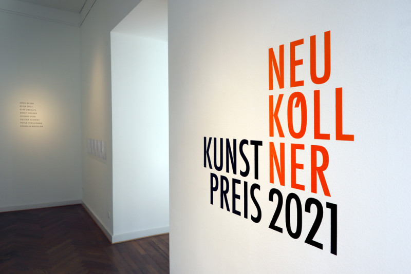 Der Titel der Ausstellung Neuköllner Kunstpreis steht in oranger Schrift auf der rechten Wand im Eingangsbereich der Galerie.