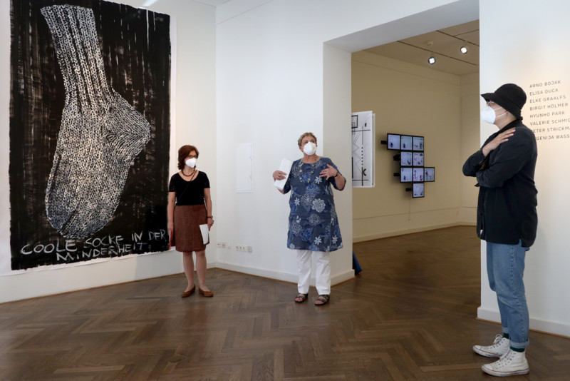 Menschen stehen in einem Halbkreis in der Galerie vor einem großformatigen Bild mit einer Socke drauf