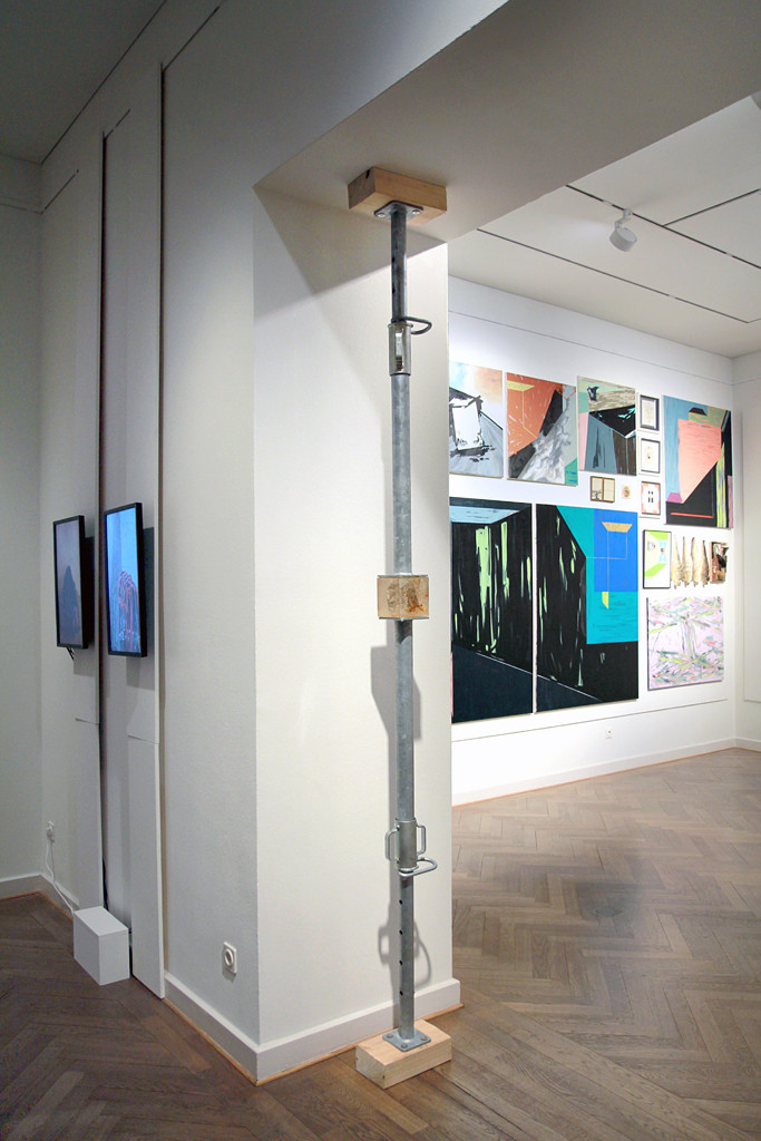In einer Schrägansicht sieht man im linken Raum zwei Bildschirme an der Wand hängen, im rechten Raum eine Wand voller abstrakter bunter Gemälde.