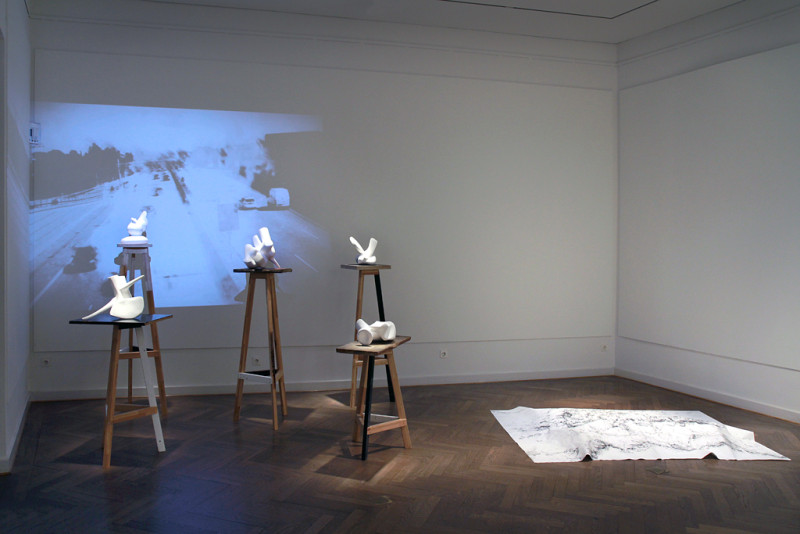 Vor einer schwarz-weißen Projektion stehen fünf Holzsockel mit weißen abstrakten Skuplturen darauf. Daneben ein großes weißes Blatt Papier auf dem Boden.