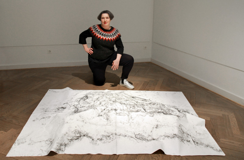 Die Künstlerin hockt auf dem Boden vor einem großen Blatt Papier mit schwarzen Graphitzeichnungen.