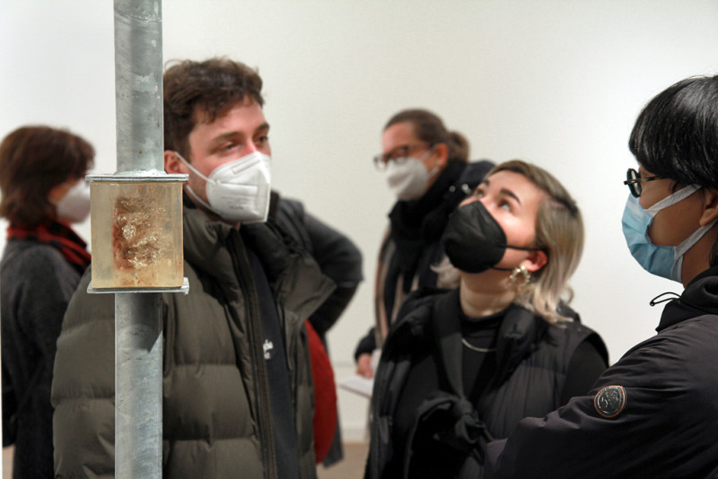 Besucher:innen mit Maske schauen sich eine Metallröhre mit einem Epoxid Quader an.