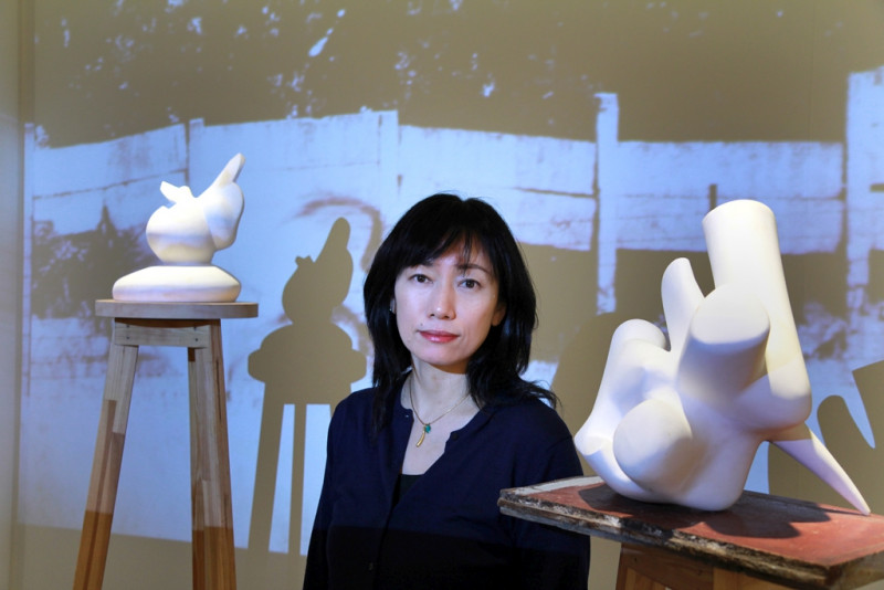 Die Künstlerin Kyoco Taniyama steht zwischen zwei Sockeln mit weißen abstrakten Objekten. Hinter ihr ist eine schwarz-weiß Projektion zu sehen.