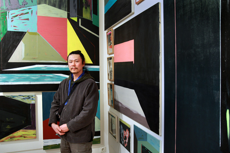 Der Künstler Bongjun Oh steht vor einer Wandgreifen Installation aus vielen bunten, abstrakt-geometrischen Gemälden.