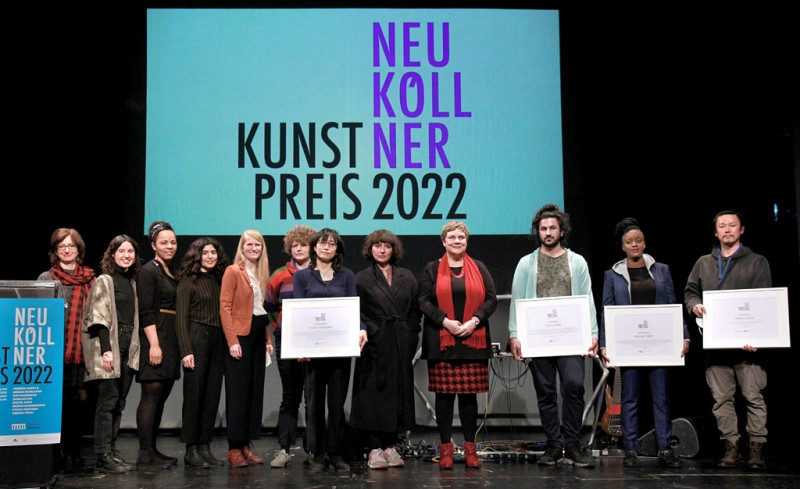 Vor dem Logo des Neuköllner Kunstpreises 2022 stehen die Preisträger:innen und Jurymitglieder mit Urkunden.